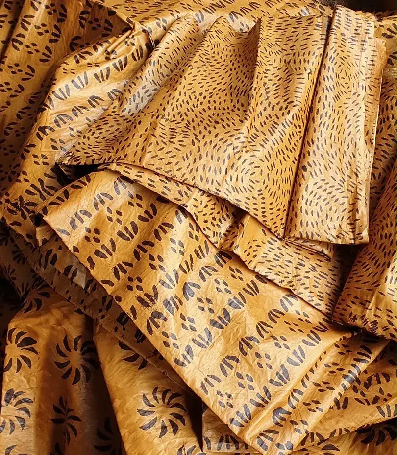 Bazin teinté jaune d’or, drapé en plis profonds, motif de graines sombres.