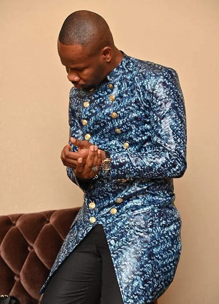Homme de profil en veste de bazin riche teinté indigo, à boutons dorés, ajustant sa montre.