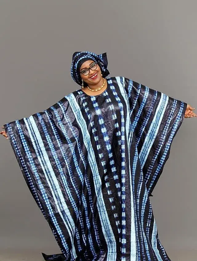 Femme présentant un boubou en bazin riche teinté indigo, bras ouverts : le tissu tombe en larges rayures bleues et blanches.